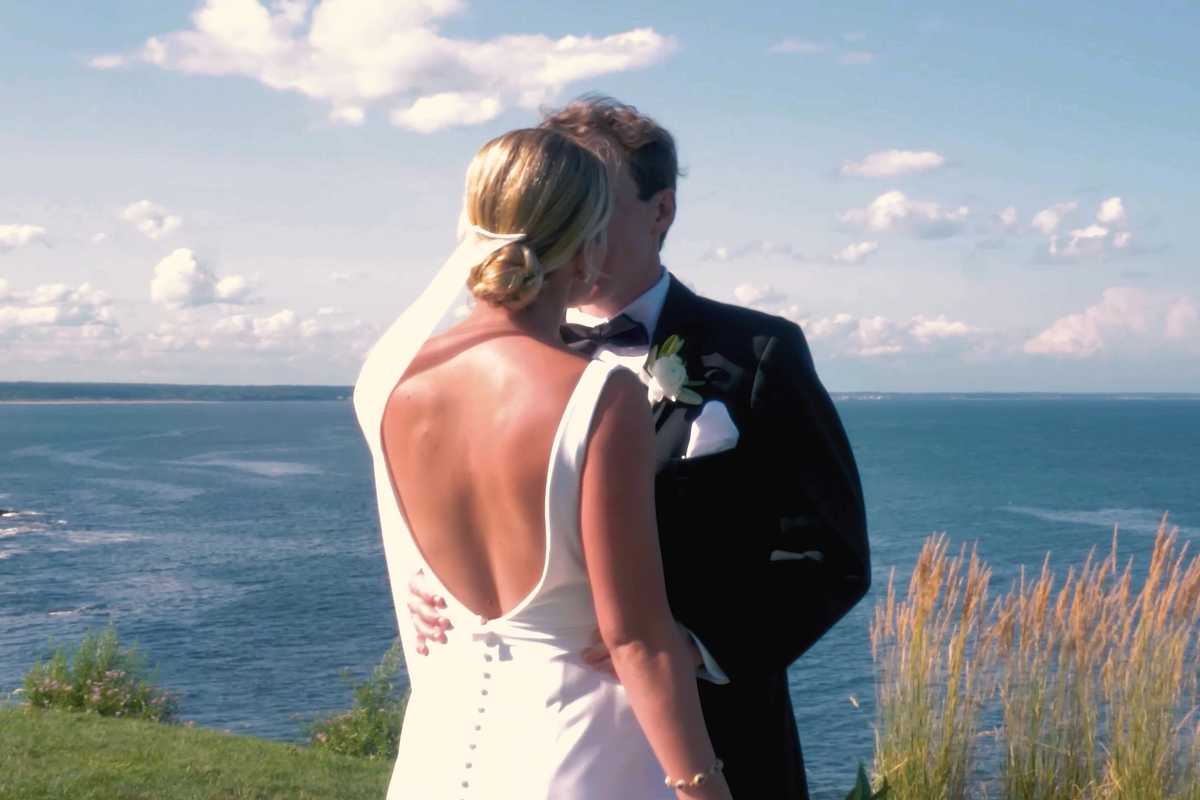 Bride and groom holding each other on a grassy cliff above the ocean, her open-back gown catching the sun