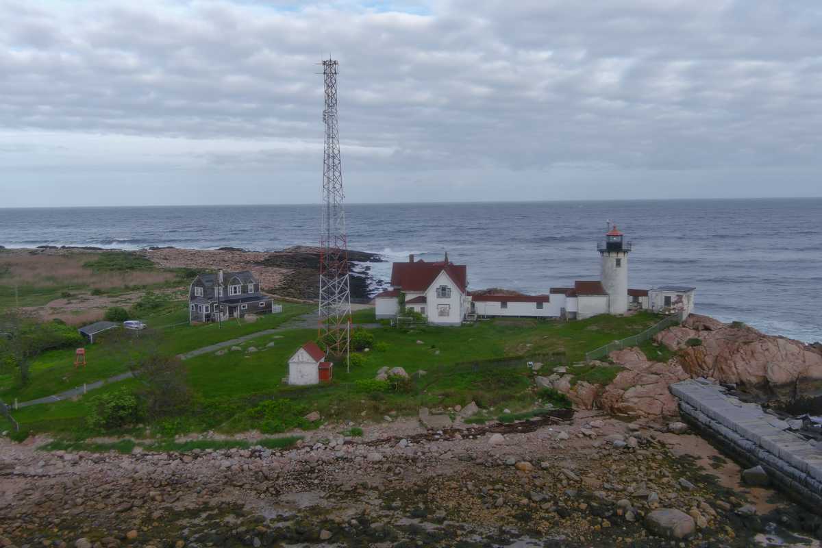 Aerial view of a white lighthouse and keeper’s house on a rocky point, waves breaking along the shore