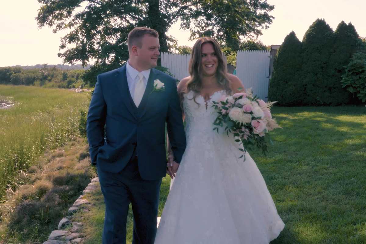 Newlyweds walking hand in hand across a waterfront lawn at golden hour, the bride carrying a blush bouquet