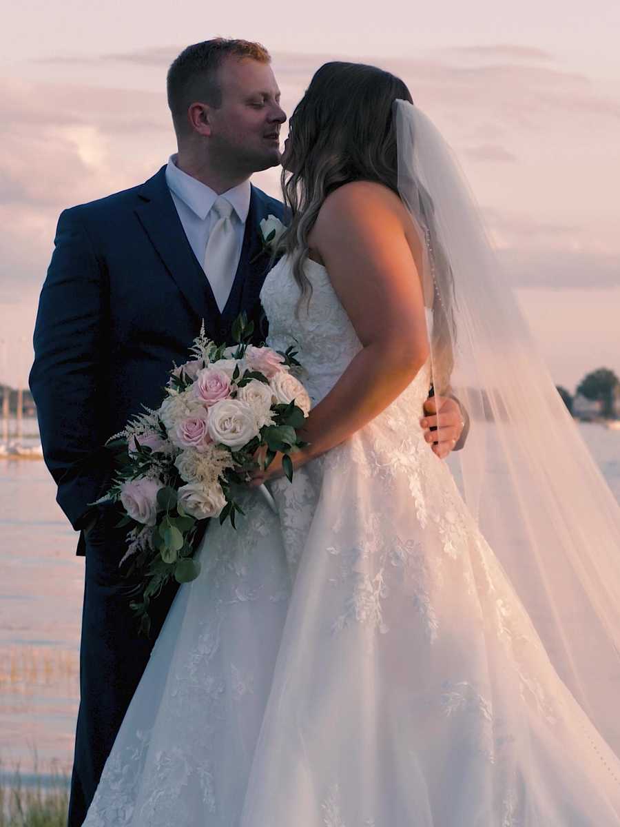 Bride and groom at the water’s edge beneath a pastel pink sunset, the bride holding a pale bouquet